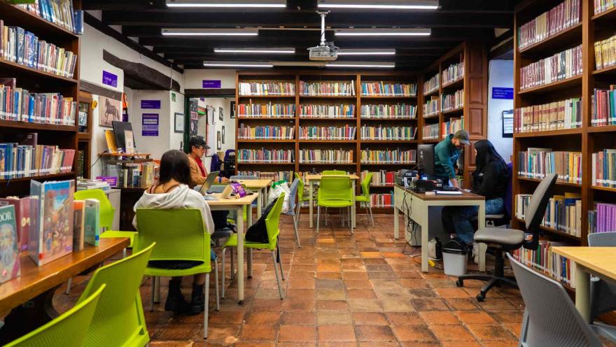 Sala de biblioteca con estanterías llenas de libros y mesas de estudio, donde varias personas leen y trabajan en un ambiente tranquilo y organizado.