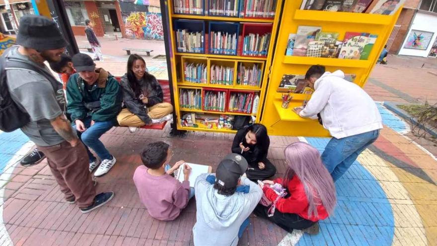 Grupo de jóvenes participa en una actividad cultural y de lectura alrededor de una biblioteca comunitaria al aire libre en Bogotá.