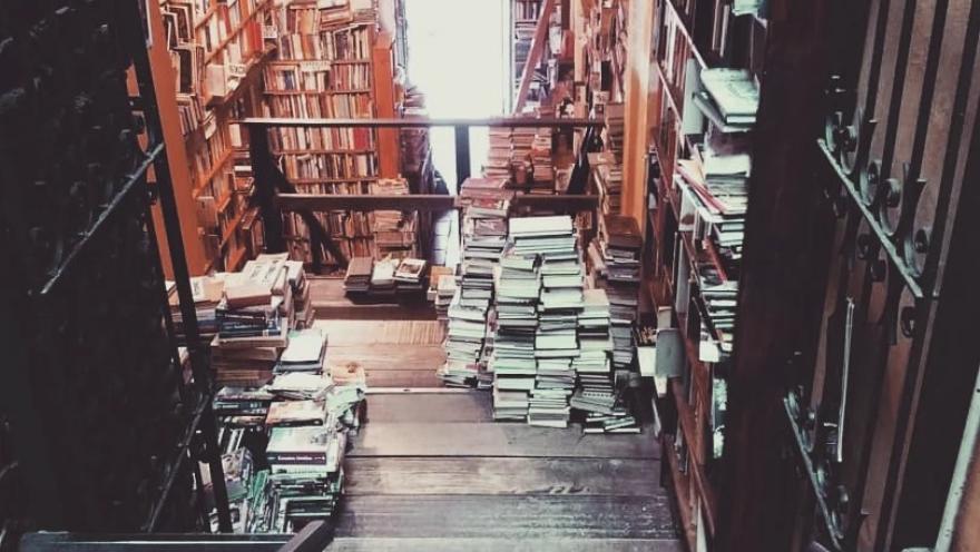  Escaleras de madera que descienden a una librería llena de estanterías y pilas de libros, iluminada por luz natural al fondo.