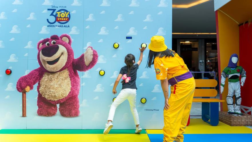 Niña participa en una actividad interactiva de Toy Story junto a una animadora en un centro comercial decorado con personajes de Disney Pixar.