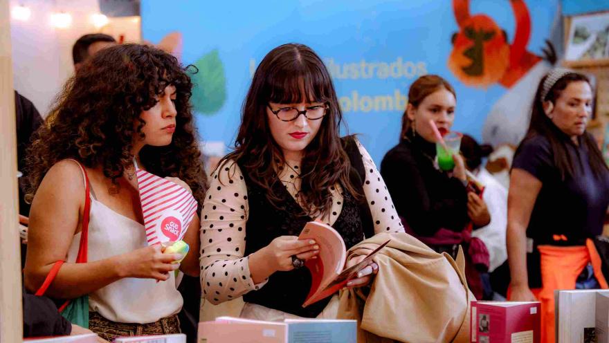 Visitantes observan y hojean libros en un stand de ilustración y literatura durante una feria cultural, en un ambiente dinámico y lleno de actividades editoriales.