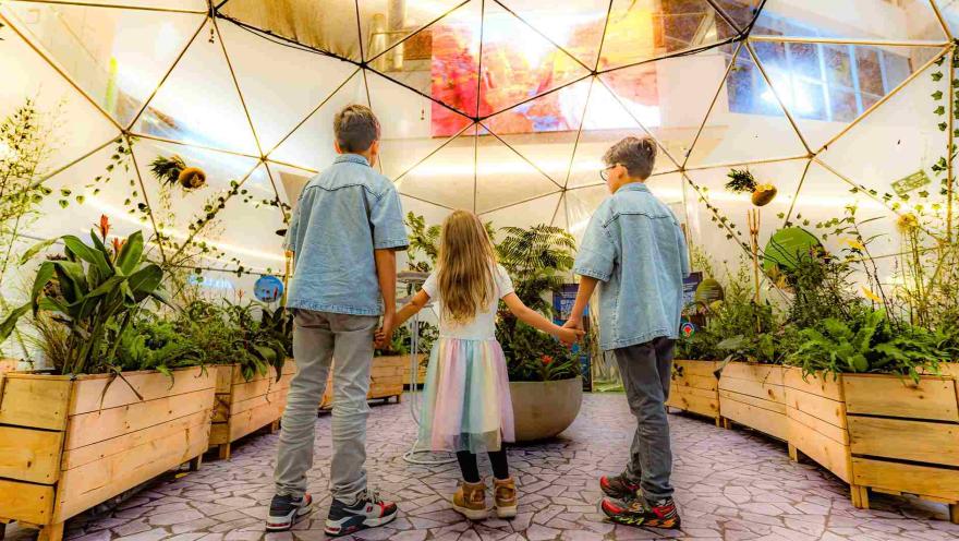 Tres niños observan una instalación interactiva rodeada de plantas dentro de una estructura tipo domo en un espacio interior.