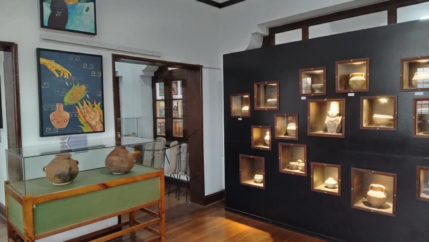 Sala de exposición de un museo arqueológico con piso de madera, vitrinas de vidrio y nichos iluminados en la pared que exhiben piezas de cerámica precolombina. En las paredes se observan pinturas de temática indígena y, al fondo, una puerta comunica con otra sala del museo.