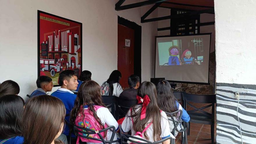 Grupo de estudiantes sentados en sillas dentro de un museo, observando una proyección en pantalla de una animación. La sala tiene paredes blancas, vigas de madera en el techo y un afiche ilustrado colgado en la pared lateral.