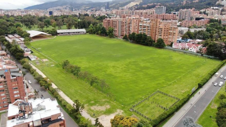Vista aérea de una amplia cancha de césped rodeada de árboles, edificios residenciales y vías urbanas, con los cerros orientales al fondo.