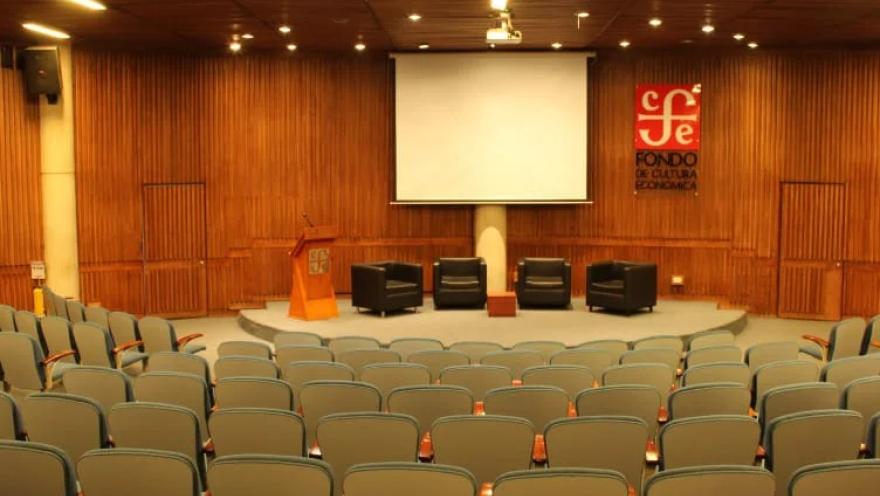 Auditorio Rogelio Salmona del Centro Cultural Gabriel García Márquez, con filas de sillas para el público frente a un escenario equipado con pantalla de proyección, atril y cuatro sillones. El recinto destaca por sus acabados en madera y su disposición para conferencias, conversatorios y actividades culturales.