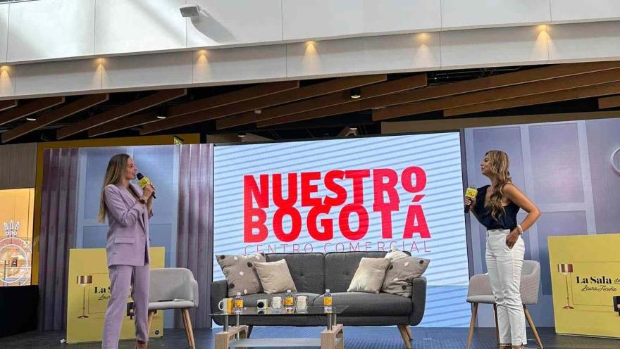 Dos mujeres participan en una charla en tarima dentro del centro comercial Nuestro Bogotá, frente a una pantalla y un público asistente.