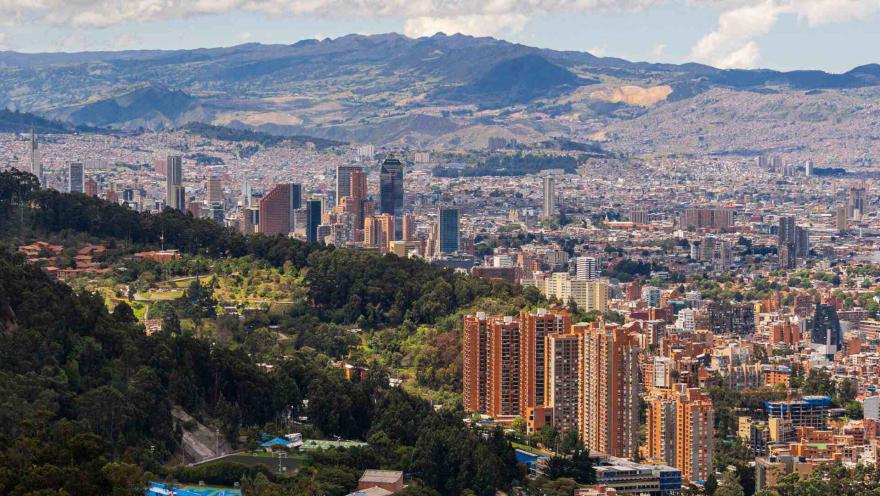 Vista panorámica de Bogotá desde los cerros orientales, con edificios, zonas verdes y montañas que rodean la ciudad bajo un cielo parcialmente nublado.