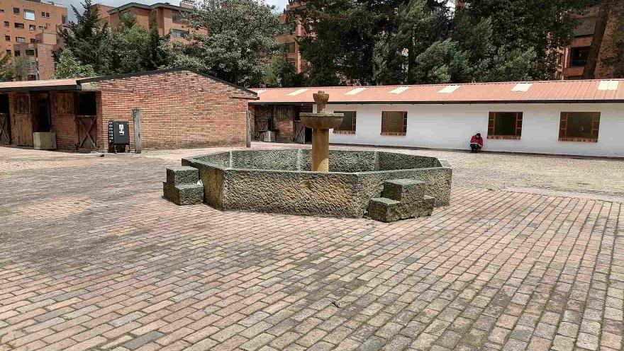 Patio adoquinado con una fuente de piedra en el centro, rodeado por edificaciones de estilo tradicional, árboles altos y edificios residenciales al fondo.