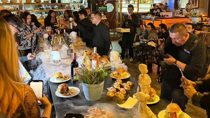 Personas participan en una experiencia gastronómica en un restaurante, mientras fotografían y disfrutan diferentes platos y bebidas sobre una mesa decorada.