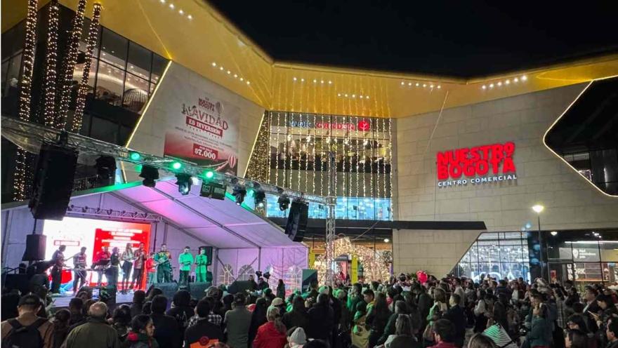  Concierto al aire libre frente al centro comercial Nuestro Bogotá, con escenario iluminado, público asistente y decoración nocturna.