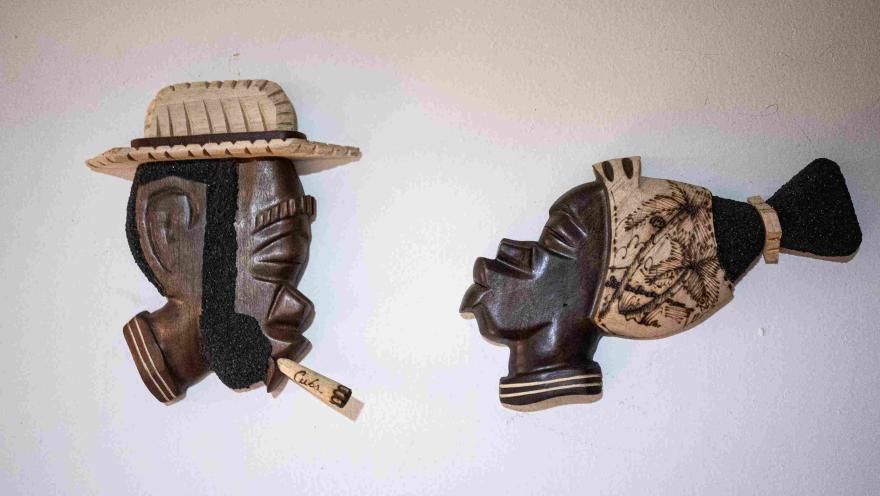 Dos máscaras artesanales de madera, decoradas con sombreros y detalles tallados, exhibidas sobre una pared blanca como parte de una colección cultural.