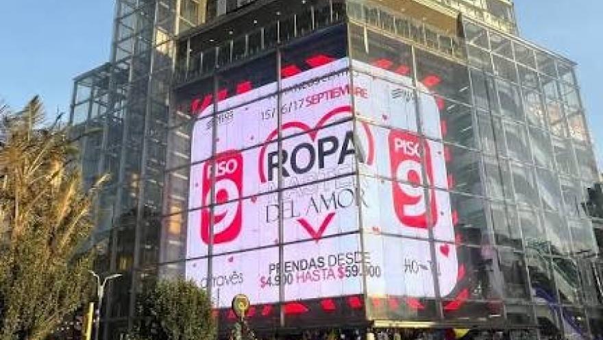 Vista exterior de un moderno edificio comercial de varios pisos con fachada de vidrio y una gran pantalla publicitaria digital. En el primer plano se observa una concurrida intersección con peatones, ciclistas y vehículos, reflejando el dinamismo del sector comercial de San Victorino, en Bogotá.
