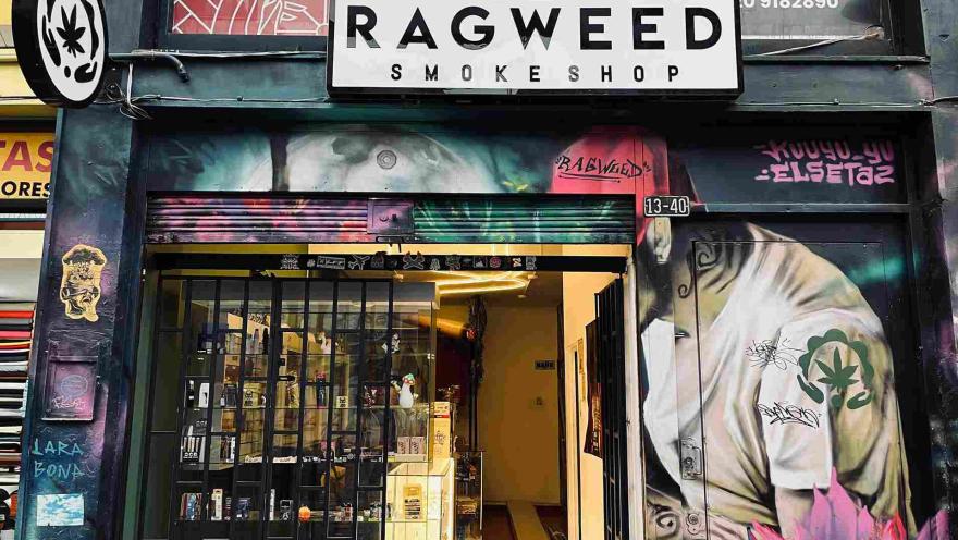 Fachada de una tienda con el letrero "Ragweed Smokeshop", rodeada de murales de arte urbano y grafitis. La entrada, con reja negra y vitrinas iluminadas, exhibe diversos productos en su interior.