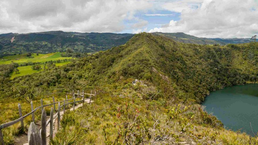 Sendero ecológico con barandas de madera entre montañas andinas, junto a una laguna de aguas verdes y un paisaje de vegetación nativa bajo un cielo parcialmente nublado.