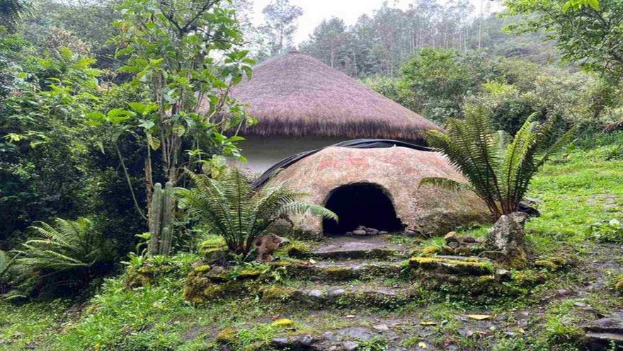 Construcción de arquitectura ancestral con techo de paja y horno de barro, rodeada de abundante vegetación y bosque nativo.