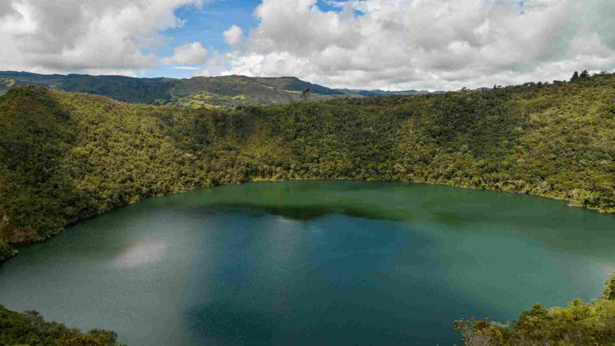Vista panorámica de la Laguna de Guatavita, rodeada de montañas cubiertas de vegetación bajo un cielo con nubes blancas. 