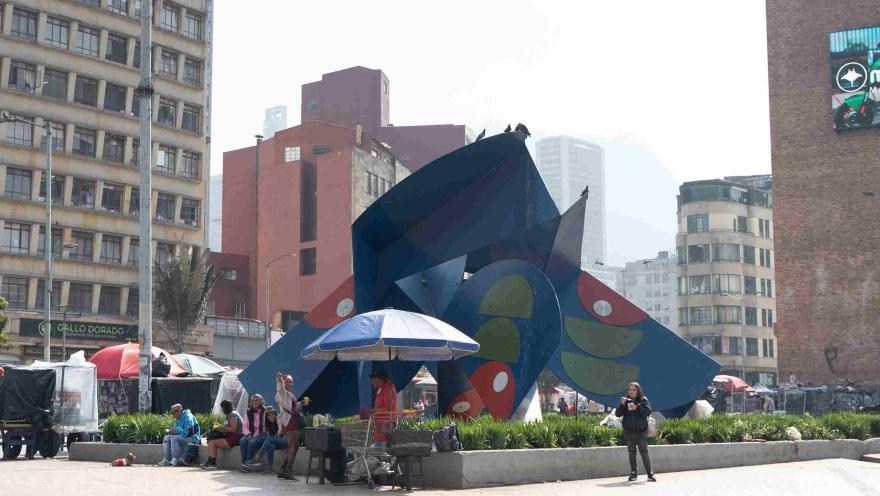 Escultura urbana de gran formato en una plaza del sector de San Victorino, rodeada de zonas verdes, vendedores y peatones. Al fondo se observan edificios del centro de Bogotá, reflejando el ambiente comercial, cultural y cotidiano de esta emblemática zona de la ciudad.