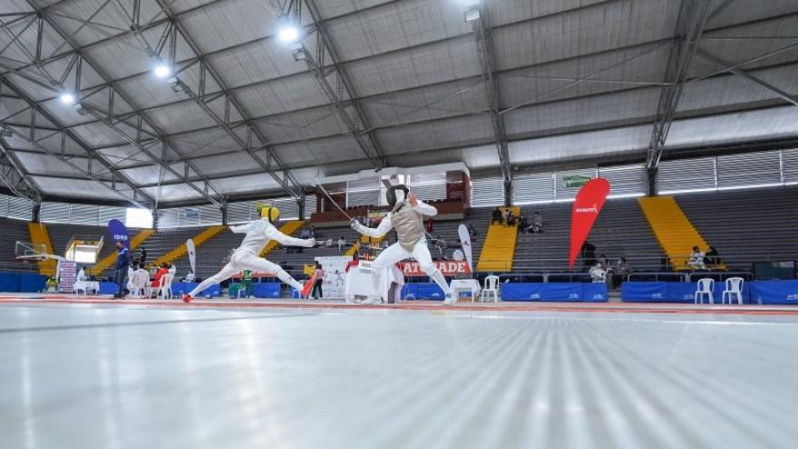 Deportistas compiten en un combate de esgrima dentro del Palacio de los Deportes de Bogotá, en una pista rodeada por graderías y escenarios de competencia.