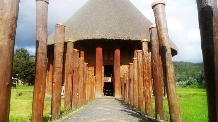 Camino de piedra rodeado de columnas de madera que conduce a un templo circular con techo de paja, en medio de un paisaje verde y bajo un cielo parcialmente nublado.
