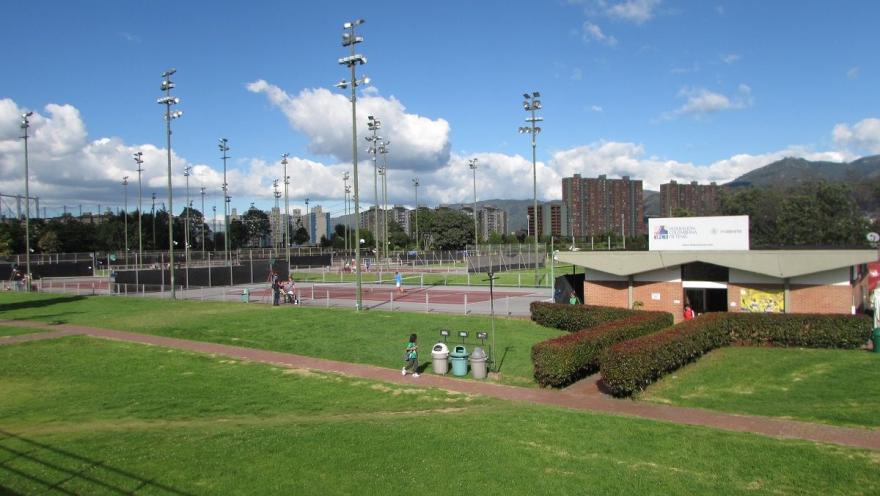 Vista de las canchas de tenis del Centro de Alto Rendimiento de Bogotá, rodeadas de zonas verdes, iluminación deportiva y edificios de la ciudad al fondo.