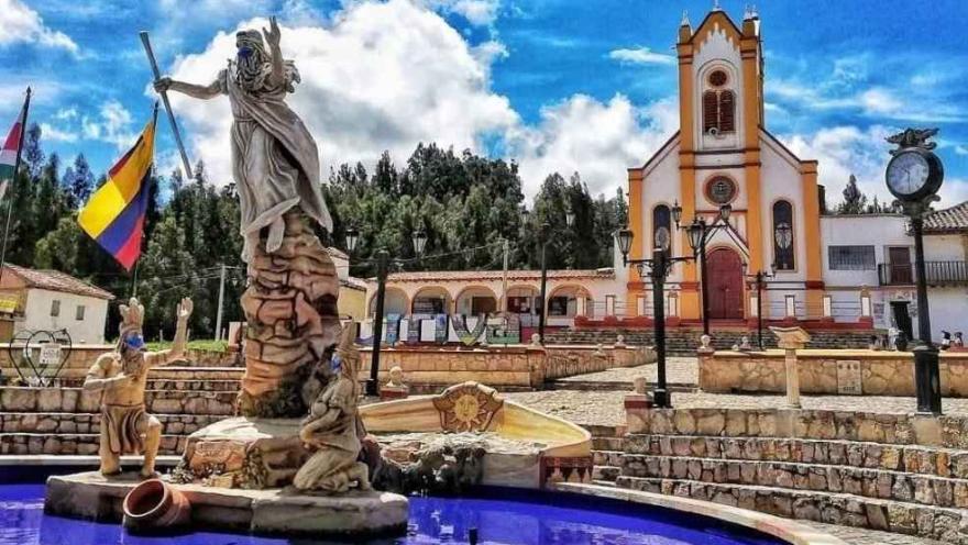 Fuente con esculturas y un monumento central en una plaza, frente a una iglesia de fachada blanca y amarilla, bajo un cielo azul con nubes.