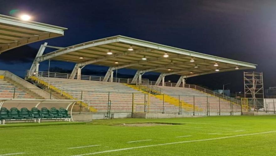  Vista del Estadio Olaya Herrera durante la noche, con la cancha iluminada, las graderías cubiertas y el banco de suplentes en primer plano.