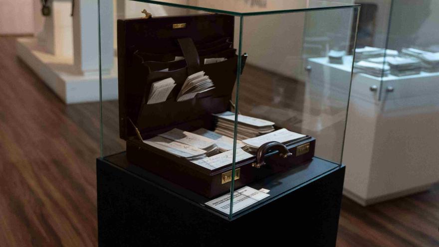  Maletín antiguo con documentos y cheques históricos, protegido por una vitrina de vidrio y exhibido como pieza museográfica.