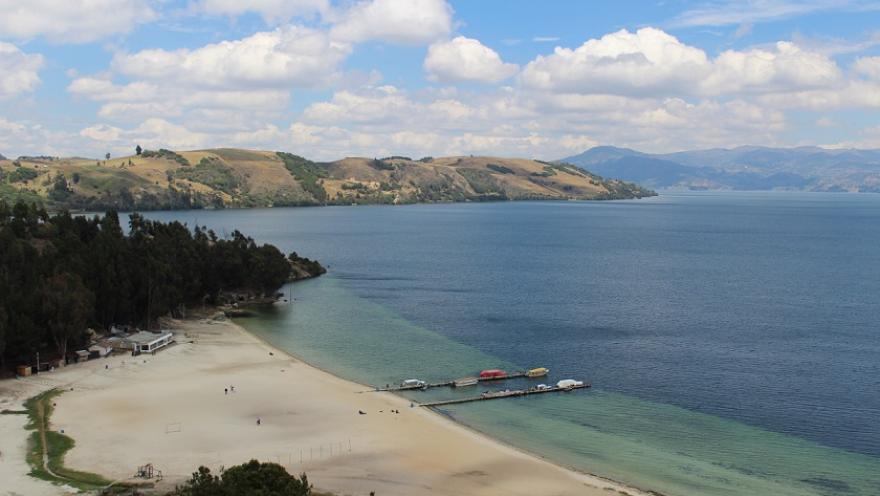 Vista panorámica de una playa de arena clara junto a un lago de aguas azules y tranquilas, con un muelle, colinas verdes y un cielo parcialmente nublado al fondo.