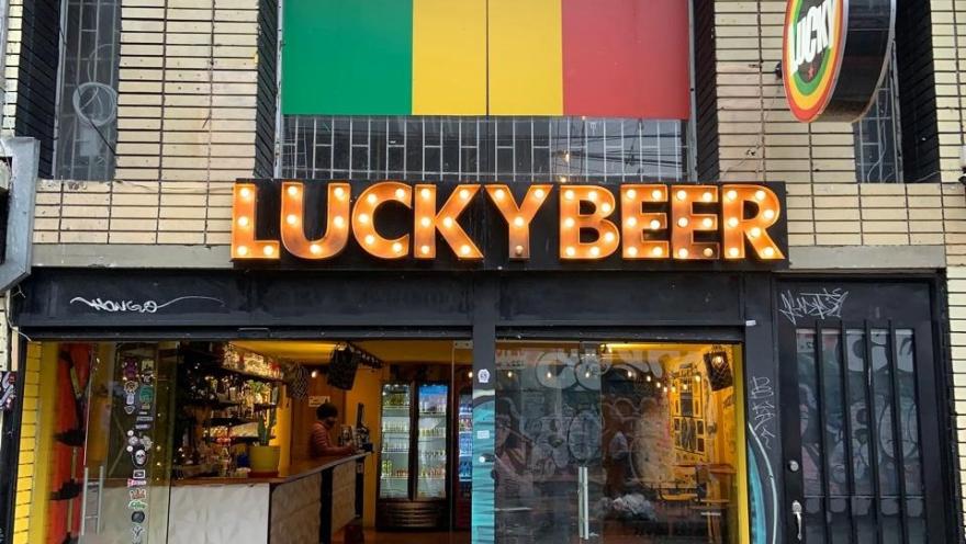 Fachada de Lucky Beer con un letrero luminoso, una bandera con colores verde, amarillo y rojo sobre la entrada, y un bar visible a través de sus puertas de vidrio.