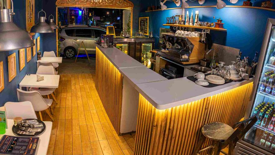  Interior de una cafetería con barra de madera iluminada, máquina de café, mesas blancas y decoración en tonos azules, vista desde la entrada durante la noche.