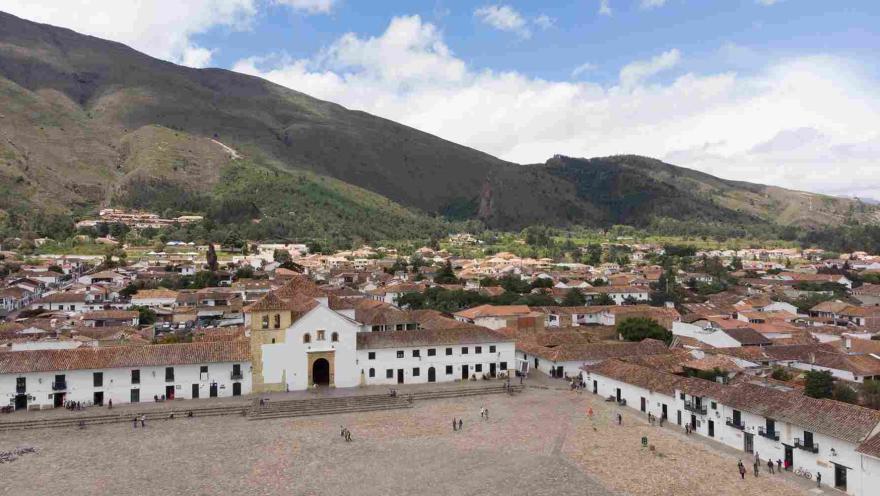 Vista panorámica de la Plaza Mayor de Villa de Leyva, rodeada de casas coloniales de techos de teja, con la iglesia principal y montañas verdes al fondo. Interior de una iglesia colonial con bancas de madera, un arco decorado con detalles dorados y rojos, y un altar principal ornamentado al fondo.