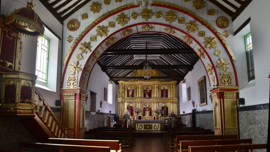 Interior de una iglesia colonial con bancas de madera, un arco decorado con detalles dorados y rojos, y un altar principal ornamentado al fondo.