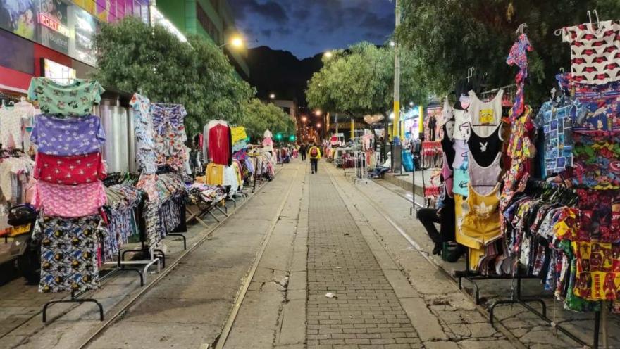 Calle peatonal de San Victorino durante una jornada nocturna de compras, con puestos de ropa y coloridas prendas exhibidas a ambos lados del camino. La iluminación y el movimiento comercial reflejan el ambiente dinámico del tradicional madrugón en el centro de Bogotá.