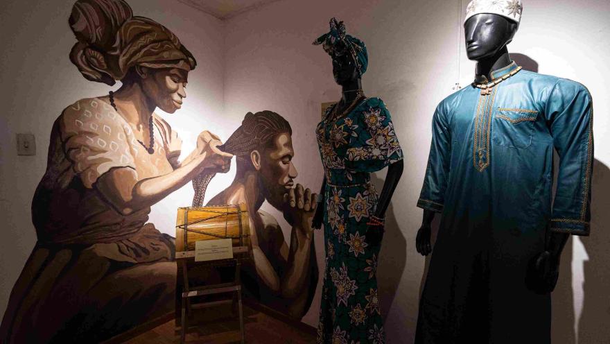 Mural de una mujer afrodescendiente trenzando el cabello de un hombre, junto a maniquíes con vestimenta tradicional africana y afrocolombiana en una exposición cultural.