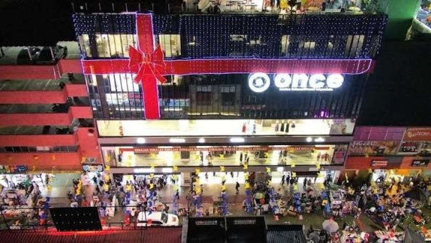 Vista nocturna del Centro Comercial Punto Once, iluminado con una llamativa decoración de luces y una gran figura de regalo en su fachada. En la calle se observa una intensa actividad comercial y numerosos visitantes, reflejando el ambiente vibrante y dinámico de San Victorino, en el centro de Bogotá.
