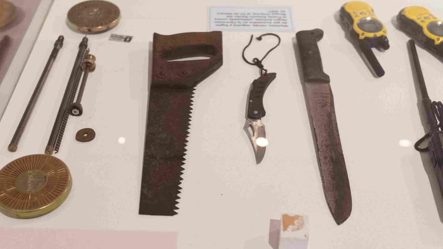 Vitrina de museo con herramientas y equipos utilizados en investigaciones forenses, entre ellos una sierra manual, un machete, una navaja y dispositivos de comunicación.