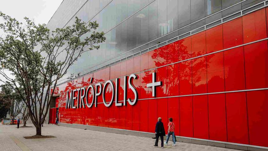 La llamativa fachada roja del Centro Comercial Metrópolis se destaca en el paisaje urbano, acompañada por amplios andenes y zonas peatonales que invitan a recorrer el sector.