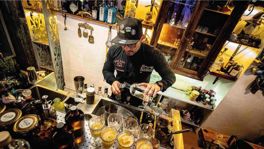 Barman con gorra sirviendo licor de una botella a un medidor sobre una barra de coctelería con vasos servidos.