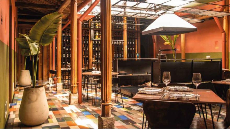 Comedor del restaurante con piso de baldosas coloridas, columnas de madera, cava de vinos y cocina abierta al fondo.