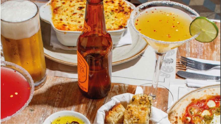 Mesa servida con cócteles, vaso de cerveza, botella de cerveza, pan artesanal, entrada horneada y pizza.