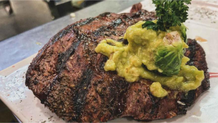 Jugoso corte de carne a la parrilla sobre plato blanco, cubierto con una porción generosa de guacamole y perejil.