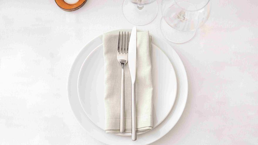 Mesa de restaurante con plato blanco, cubiertos y servilleta, preparada para recibir a los comensales.