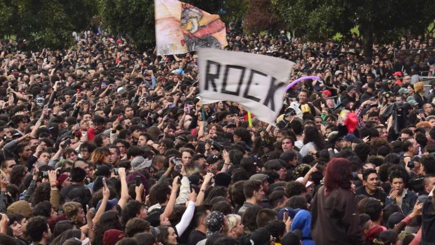Multitud de personas en un concierto al aire libre, levantando manos y banderas, con un cartel que dice “ROCK” en medio del público.