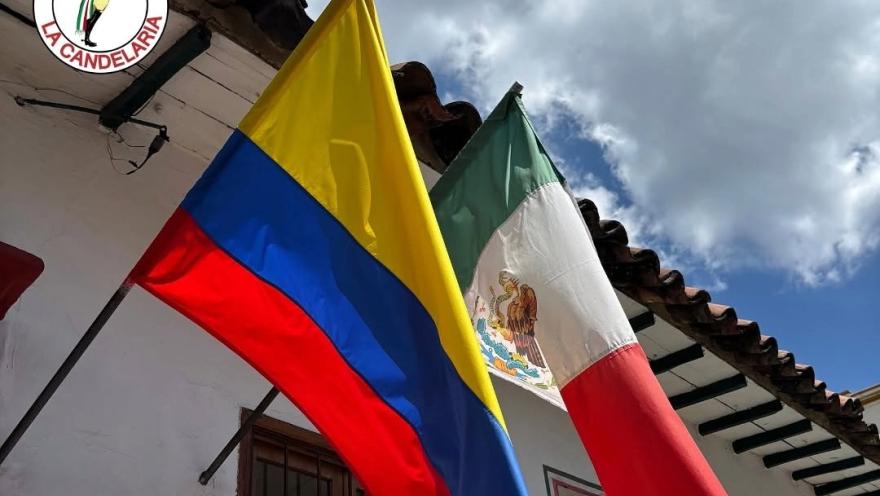 Colombian and Mexican flags wave in front of the colonial facade of Enchiladas La Candelaria under a partly cloudy sky.
