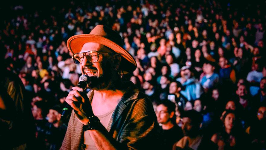 Cantante con sombrero y gafas interpreta con micrófono frente a una multitud en un concierto nocturno.