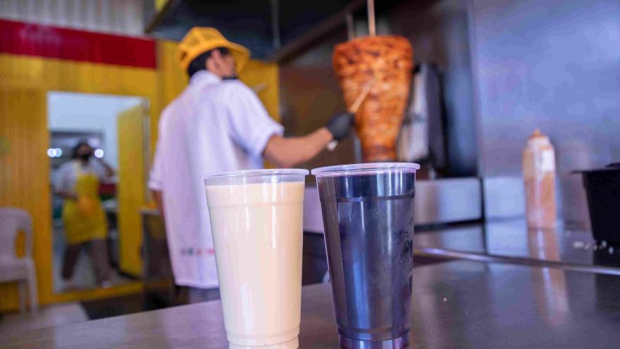  Dos bebidas servidas en vasos y un trompo de carne al pastor al fondo del restaurante.