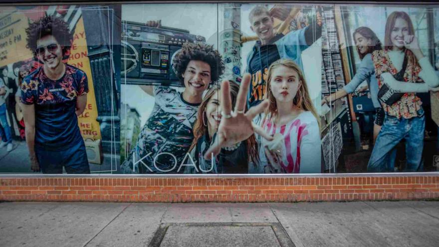 Un gran mural publicitario ocupa el escaparate de la tienda KOAJ, con imágenes de jóvenes y una estética urbana y colorida que refleja el espíritu moderno de la marca. La fachada combina los ventanales con una base de ladrillo característica de la edificación.