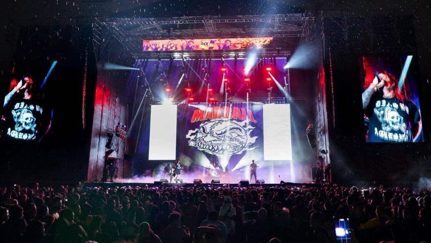 Escenario de concierto con banda de rock en vivo, luces intensas y gran público frente a la tarima durante un evento nocturno.