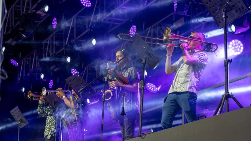 Músicos en escenario tocan trombón y saxofón bajo luces moradas durante un concierto en vivo.